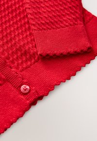 Rdeč pleteni kardigan z vzorcem teksture, rebrastimi manšetami in valovitim robom. Na sprednji strani je roza gumb za zapenjanje.