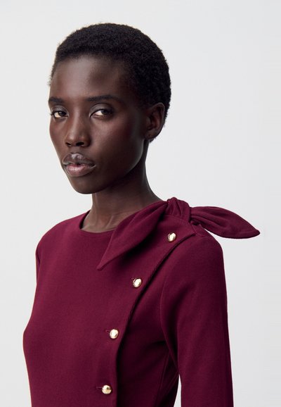 Top bordeaux con scollatura rotonda, texture a coste, bottoni laterali e un dettaglio a fiocco legato su una spalla. Accenti dorati vicino ai bottoni.