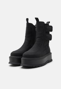 Botas negras hasta el tobillo con un diseño estriado, cierre de cremallera y correas de Velcro. Fabricadas en material sintético con una suela de plataforma gruesa.