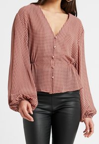 Halenka s dlouhým rukávem a knoflíkovým zapínáním v růžovém houndstooth vzoru. Vybavena hlubokým výstřihem do V a nařasenými manžetami, kombinována s vypasovanými černými kalhotami.
