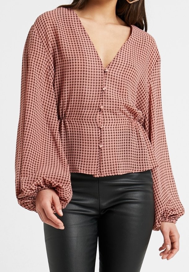Halenka s dlouhým rukávem a knoflíkovým zapínáním v růžovém houndstooth vzoru. Vybavena hlubokým výstřihem do V a nařasenými manžetami, kombinována s vypasovanými černými kalhotami.