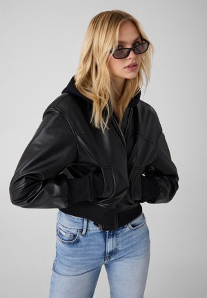 Blonde Frau trägt schwarze Lederjacke und Sonnenbrille, steht mit den Händen in den Jackentaschen vor einem einfarbigen grauen Hintergrund.