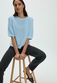 Vaaleansininen neulepaita lyhyillä kellotuloilla, yhdistetty tummiin farkkuihin. Malli istuu puupenkillä, kädet polvilla.