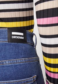 Jeans de ganga com um tom escuro e costuras visíveis, apresentando uma etiqueta preta da DRDENIM na cintura, combinados com uma blusa de manga longa listrada.