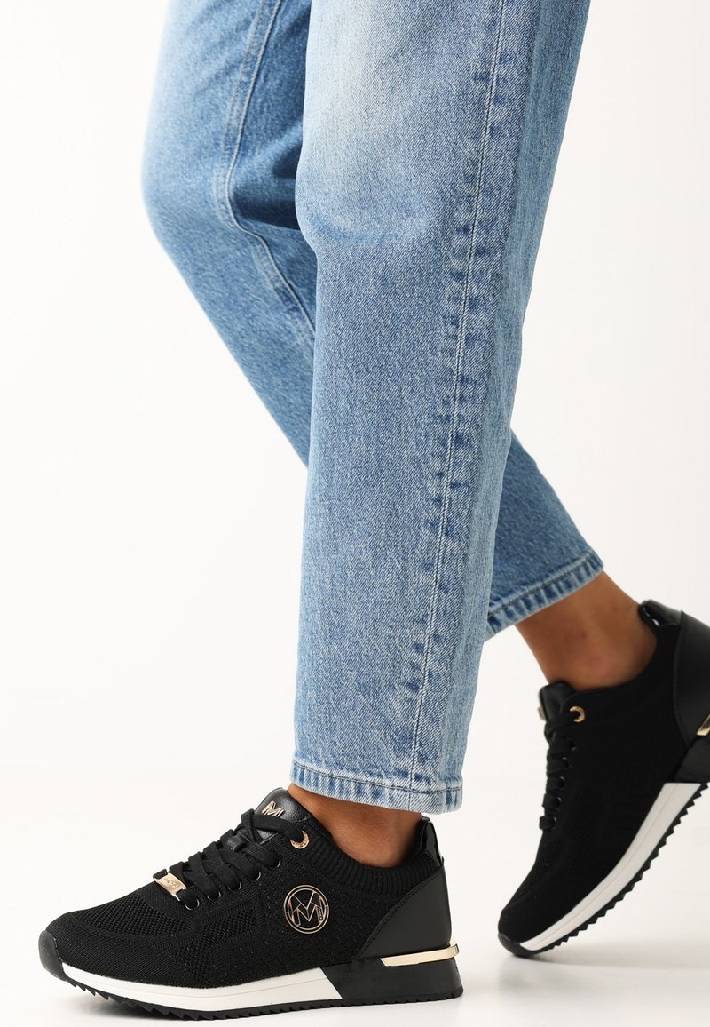 Schwarze Strick-Sneaker mit goldenen Akzenten, ausgestattet mit einem strukturierten Obermaterial, eleganter Form und weißer Sohle, kombiniert mit hellblauen, hochgekrempelten Jeans.