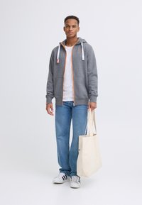 Moški v sivi jopa z zadrgo, beli majici, modrih jeansh in belih supergah, ki drži bež torbo na enobarvni podlagi.