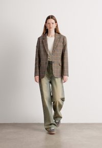 Blazer à carreaux marron, avec deux boutons, associé à un pantalon vert pâle et ample, et des chaussures grises. Texture lisse avec une coupe décontractée.