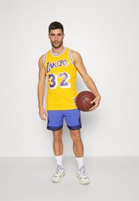 Canottiera da basket gialla con la scritta "Lakers" e numero 32, abbinata a pantaloni blu con strisce nere, mentre tiene in mano un pallone da basket.