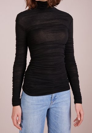 Personne portant un haut à col roulé noir à manches longues froncées et un jean denim bleu clair sur un fond uni.