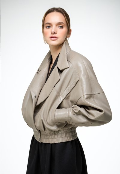 Jeune femme aux cheveux blonds portant une veste en cuir taupe oversized par-dessus une robe noire, posant devant un fond blanc.