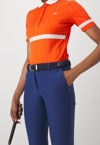 Orange golfpolo med ribbad struktur och vit horisontell rand; kombinerad med marinblå byxor och ett flätat bälte. Detaljer inkluderar krage och logotyp.