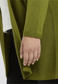 Cárdigan acanalado en verde oliva con un diseño en capas. Primer plano de una mano agarrando la tela en el dobladillo, mostrando la textura y el detalle.