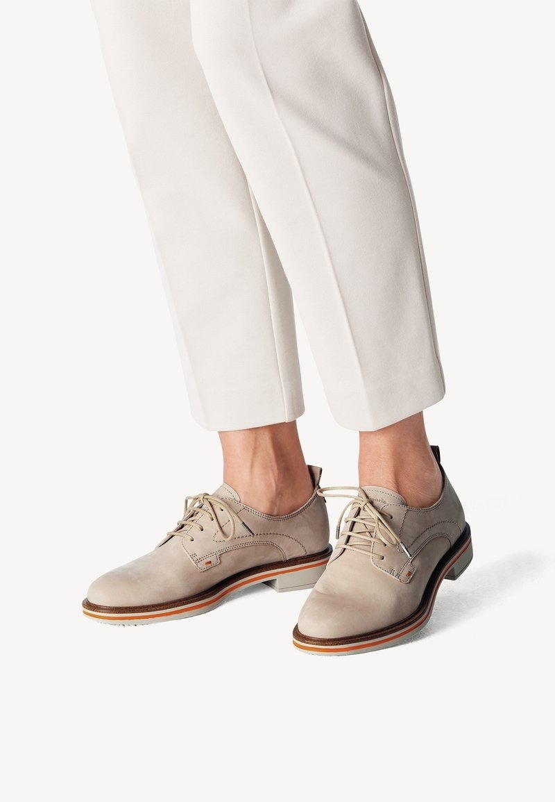 Chaussures à lacets en cuir beige portées avec un pantalon blanc raccourci, montrant des coutures détaillées et un design de semelle à couches.