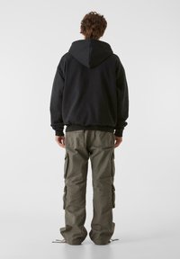 Fekete kapucnis pulóver laza fitettel, olíva zöld cargo nadrággal párosítva, amely több zsebet tartalmaz és tapered fazonú.