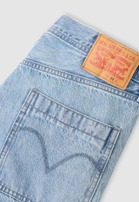 Ανοιχτό μπλε τζιν, με καφέ δερμάτινο ταμπελάκι με κόκκινη εκτύπωση, αντίθετες ραφές και χαρακτηριστικό σχέδιο πίσω τσέπης.