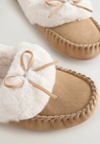 Tan suede slippers with a fluffy white faux fur cuff, finished with a bow and hand-stitched detailing along the edges. Soft-textured surface.