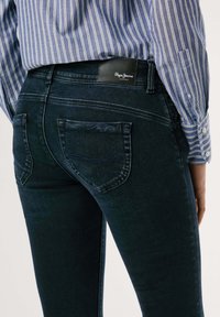 Temno modri denim kavbojki z ozkim krojem, ki imajo zadnje žepe in subtilne šive. Pas ima črno oznako z logotipom blagovne znamke.