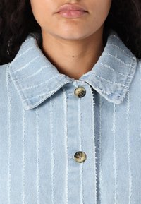 Ljusblå denimskjorta med randigt mönster, med klassisk krage och två bruna knappar. Tyget har en fransig textur.