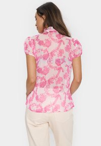 Permatoma rožinė palaidinė su gėlėta raštu, trumpomis pūstomis rankovėmis, raukšlėtu kaklu ir prigludusia liemeniu. Nešiojama su šviesiomis kelnėmis.