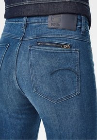 Gros plan sur un jean denim bleu avec une poche arrière comportant une fermeture éclair horizontale et un patch de marque en cuir sur la ceinture.