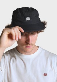 Čierny klobúk s vlekom vyrobený z textúrovanej tkaniny, s námestným nášivkom a bielym logom slona. Nosí sa mierne naklonený, v kombinácii s bielym tričkom.