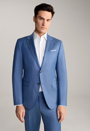 A man stands confidently in a tailored blue suit with a white shirt, against a plain gray backdrop, showcasing a modern, professional look.