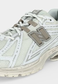 Ανοιχτό γκρι αθλητικό παπούτσι New Balance με πλέγμα πάνω μέρος, μπεζ μπαλώματα με το λογότυπο "N", λευκά κορδόνια και υφή στην σόλα με λεπτομέρειες απορρόφησης.