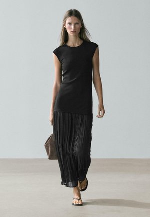 Kvinde går iført en ærmeløs sort top, en flydende sort plisseret nederdel, sorte sandaler og bærer en brun taske mod en ensfarvet baggrund.