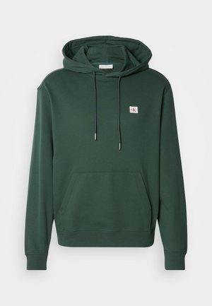 Mørkegrønn hettegenser laget av myk bomull, med en frontlomme, justerbare snor, og et lite logo-lapp på brystet.