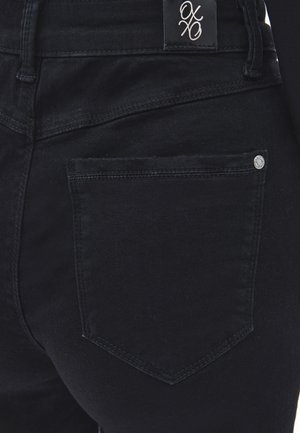 Bolsillo trasero de jeans de mezclilla negra con detalle de remache plateado y parche de cinturón negro con el logotipo estilizado "oxo" en blanco.