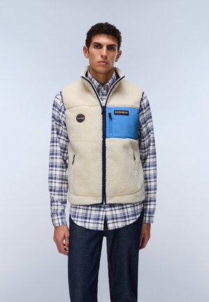 Young man wearing a cream fleece vest with a blue chest pocket over a blue and white plaid shirt and dark jeans standing against a plain background.