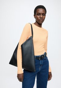 Čierna kožená taška typu tote s trojuholníkovým tvarom, hladkou textúrou a dlhým remienkom, doplnená o rebrovaný broskyňový top a vysoko pásové modré džínsy.