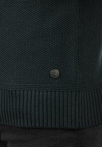 Suéter de punto verde oscuro con un cuerpo texturizado, dobladillo acanalado y un pequeño logotipo metálico en el lado inferior.