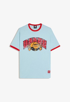 Tricou albastru deschis cu guler roșu și margini roșii la mâneci, având un design circular uzat în roșu și galben cu text și o mică etichetă roșie cu logo la tiv.