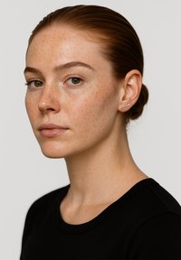 Giovane donna con i capelli rossi raccolti, leggeri nei, che indossa una maglietta nera e un piccolo orecchino in argento, guarda con calma verso la macchina fotografica.