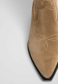 Karmelowe zamszowe botki sięgające kostki z ostrym noskiem, ozdobnym szyciem w kształcie kokardy oraz czarną gumową podeszwą. Gładka faktura.