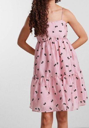 Frau trägt ein ärmelloses pinkfarbenes Kleid mit dünnen Trägern und kleinen schwarzen Blumenmustern, steht mit den Händen hinter dem Rücken.