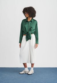 Femme aux cheveux bouclés portant des lunettes, une chemise en satin verte nouée, des culottes plissées blanches, des chaussettes blanches et des baskets blanches, debout sur un tapis bleu.