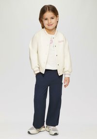 Witte bomberjack met drukknoopjes, geborduurd "Princess"-tekst, gecombineerd met marineblauwe cargo-broek en grijze sneakers. Glad stoftextuur.