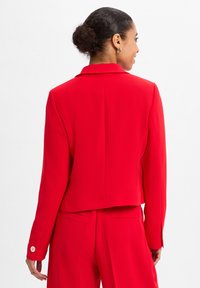 Roter Blazer mit tailliertem Design, glatter Textur, Reverskragen und Knopfakzenten an den Ärmeln. Eigenschaften sind eine verkürzte Länge und versteckte Taschen.