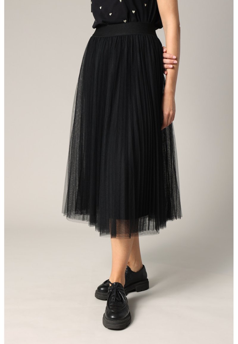 Femme portant une jupe midi plissée noire transparente avec des chaussures noires épaisses à lacets, debout sur un fond clair.