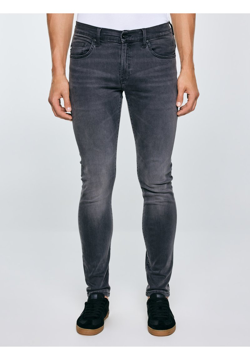 Egy férfi vékony szabású, kifakult fekete farmert és fekete, gumitalpú sportcipőt visel, egyszínű, világos háttér előtt áll.