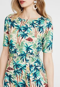 Žena v šatech s páskem, které mají tisk palmových stromů v tyrkysové a modré barvě, s krátkými rukávy a velkými kruhovými náušnicemi.