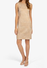 Robe beige sans manches avec un motif de feuilles subtil, coupe ajustée et longueur jusqu'aux genoux. Associée à des sandales blanches simples à lanières.