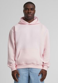Rosa hettegenser med frontlomme og snorhette, som har en liten hvit logo. Laget av myk stoff, med ribbet mansjetter og nederkant.