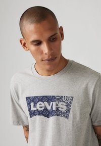 Pilkas medvilnės marškinėliai su dideliu tamsiai mėlynu Levi's logotipu, dekoruotu gėlėmis. Apvalus iškirpimas ir trumpomis rankovėmis. Kasdienis laisvas stilius.