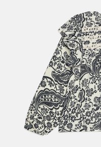 Hvit bluse med svarte blomster- og paisleymønstre, med en volangkrage og samlede ermer for ekstra tekstur og detaljer.