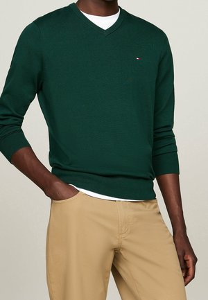 Mann trägt dunkelgrünen V-Ausschnitt-Pullover über weißem Hemd mit beigen Hosen, eine Hand in der Tasche, vor neutralem Hintergrund.