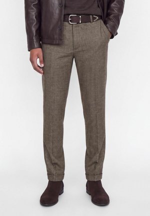 Homme portant une veste en cuir marron, une ceinture marron foncé, un pantalon sur mesure vert olive et des bottines en daim marron foncé.