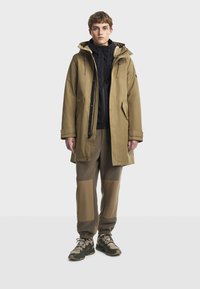 Svetlohnedá parka s kapucňou a zipsovým uzáverom, položená na čiernom topu so zipsom. Textúrované hnedé nohavice s farebnými blokmi a tmavé tenisky.